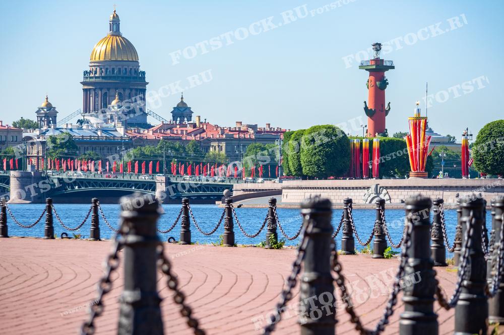Фотография демонстрирует классический вид на Исаакиевский собор и Ростральные колонны в Санкт-Петербурге. Это изображение сделано с набережной, где в кадр попали и элементы городского ограждения — чугунные столбы, соединённые цепями. Такой вид может использоваться для иллюстрации путеводителей, образовательных статей, туристических брошюр, а также в качестве красочного фона на сайтах, посвящённых Санкт-Петербургу.
